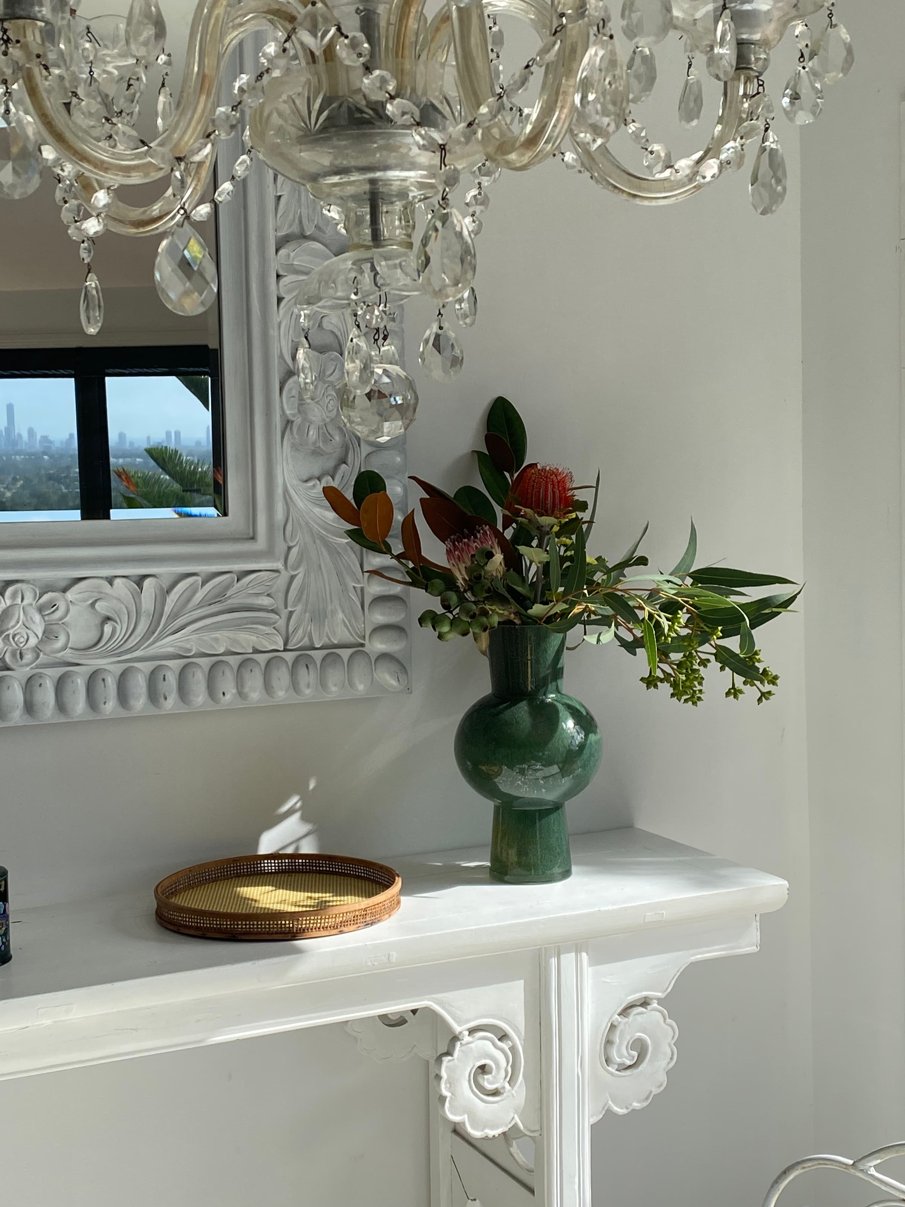 Native floral arrangement in a green vase on a white mantelpiece under a crystal chandelier.