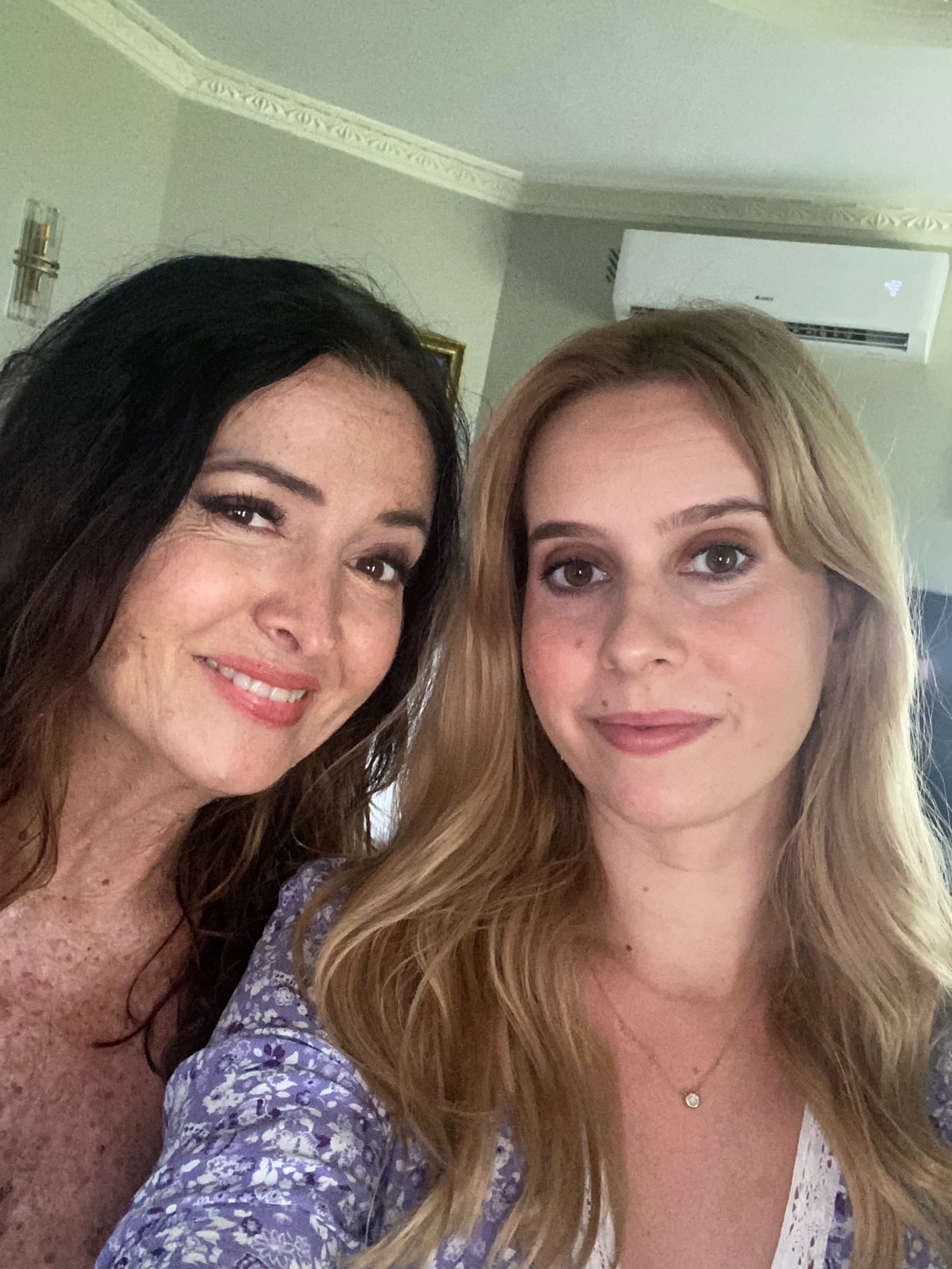 A dark-haired woman and a blonde woman smile together for a close-up indoor selfie.